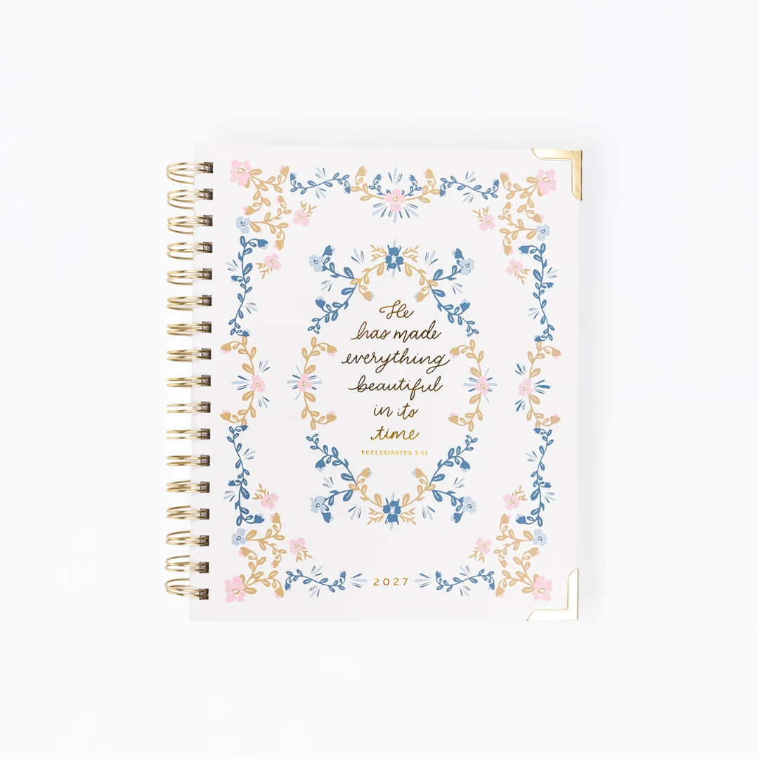 A spiral-bound 2027 planner on a clean white background. The cover features a delicate floral wreath border in shades of blue, pink, and gold, surrounding elegant gold script text that reads 'He has made everything beautiful in to June' and 'WELL DESIGNED 2027'.