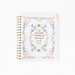 A spiral-bound 2027 planner on a clean white background. The cover features a delicate floral wreath border in shades of blue, pink, and gold, surrounding elegant gold script text that reads 'He has made everything beautiful in to June' and 'WELL DESIGNED 2027'.