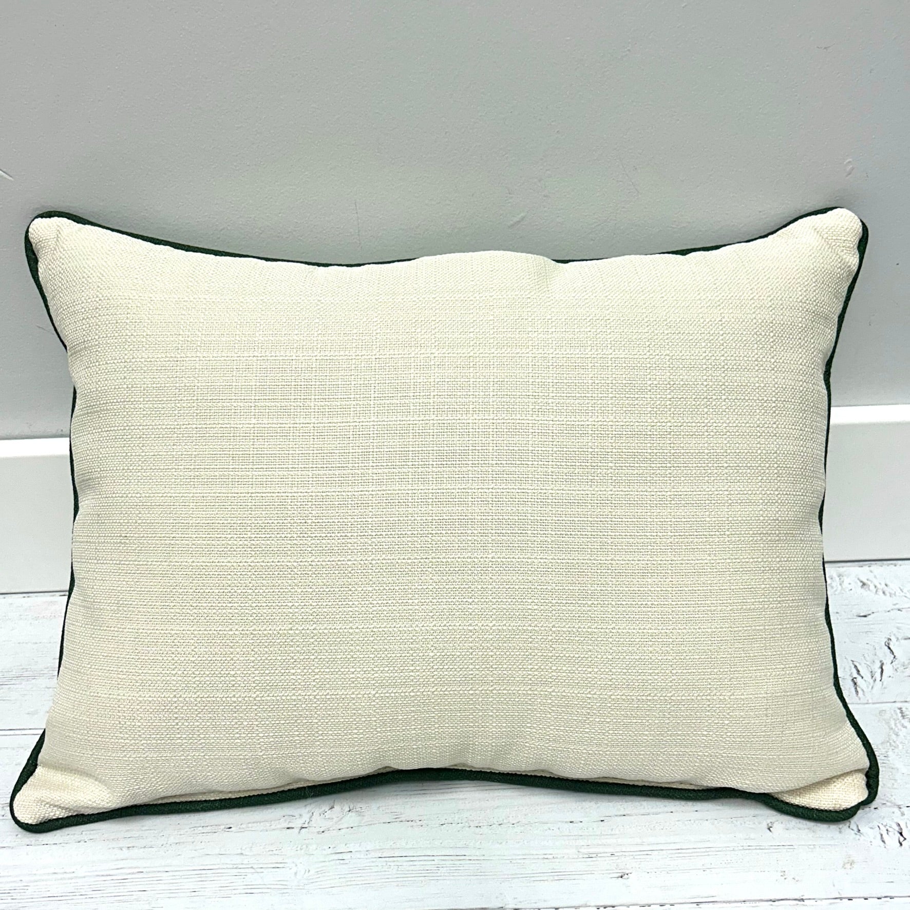 A square throw pillow with a textured cream linen-like fabric and dark green piping, resting on a white wooden floor against a gray wall.