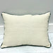A square throw pillow with a textured cream linen-like fabric and dark green piping, resting on a white wooden floor against a gray wall.