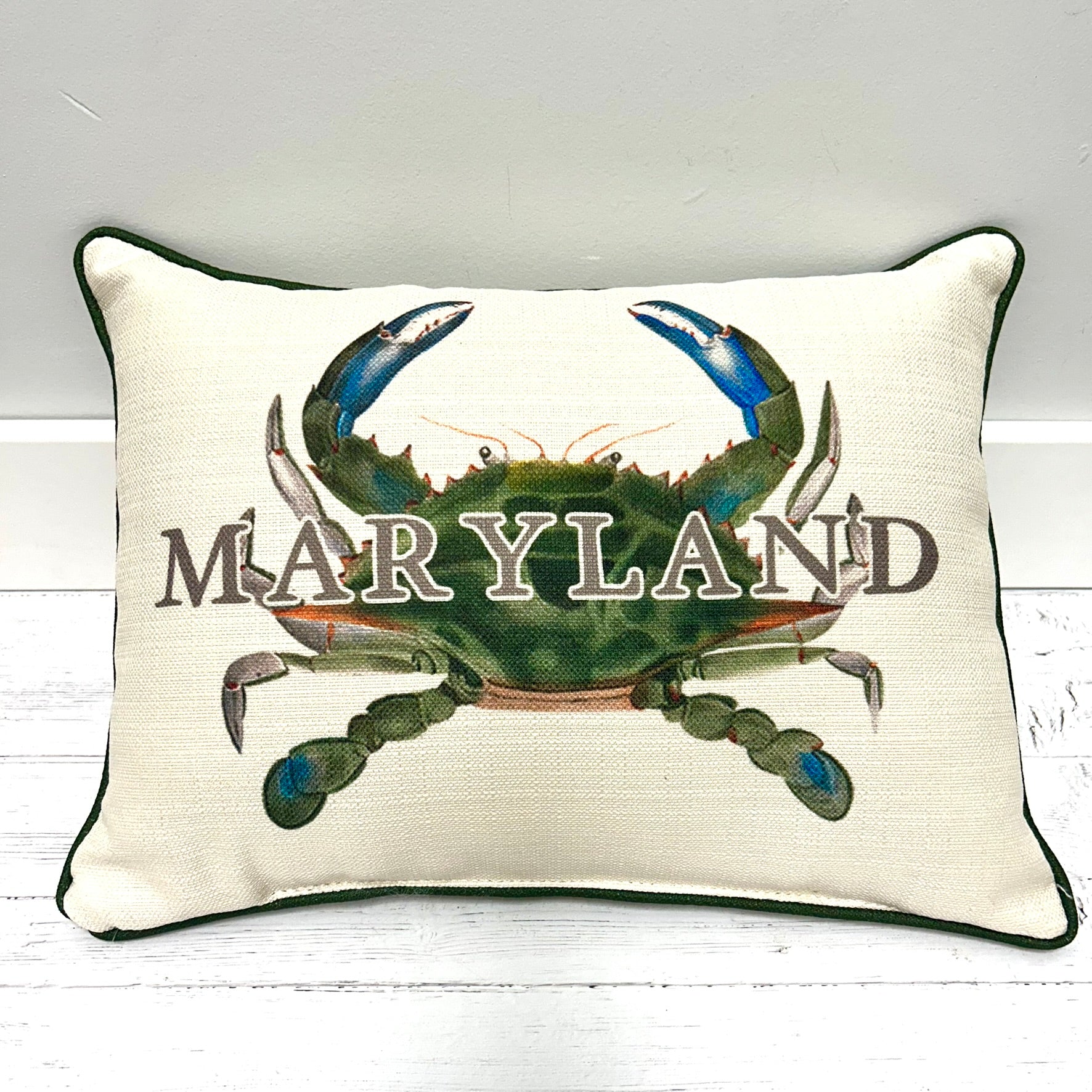 A square decorative throw pillow with a cream linen-textured background, dark green piping, and a large printed blue crab with the word 'MARYLAND' across the front, shown against a white wood surface and light wall.