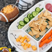 A football-themed divided snack platter on a granite countertop, filled with carrot sticks, pita chips, cheese cubes, sliced cucumbers, and salami, alongside a football-shaped dip bowl and a helmet mug.