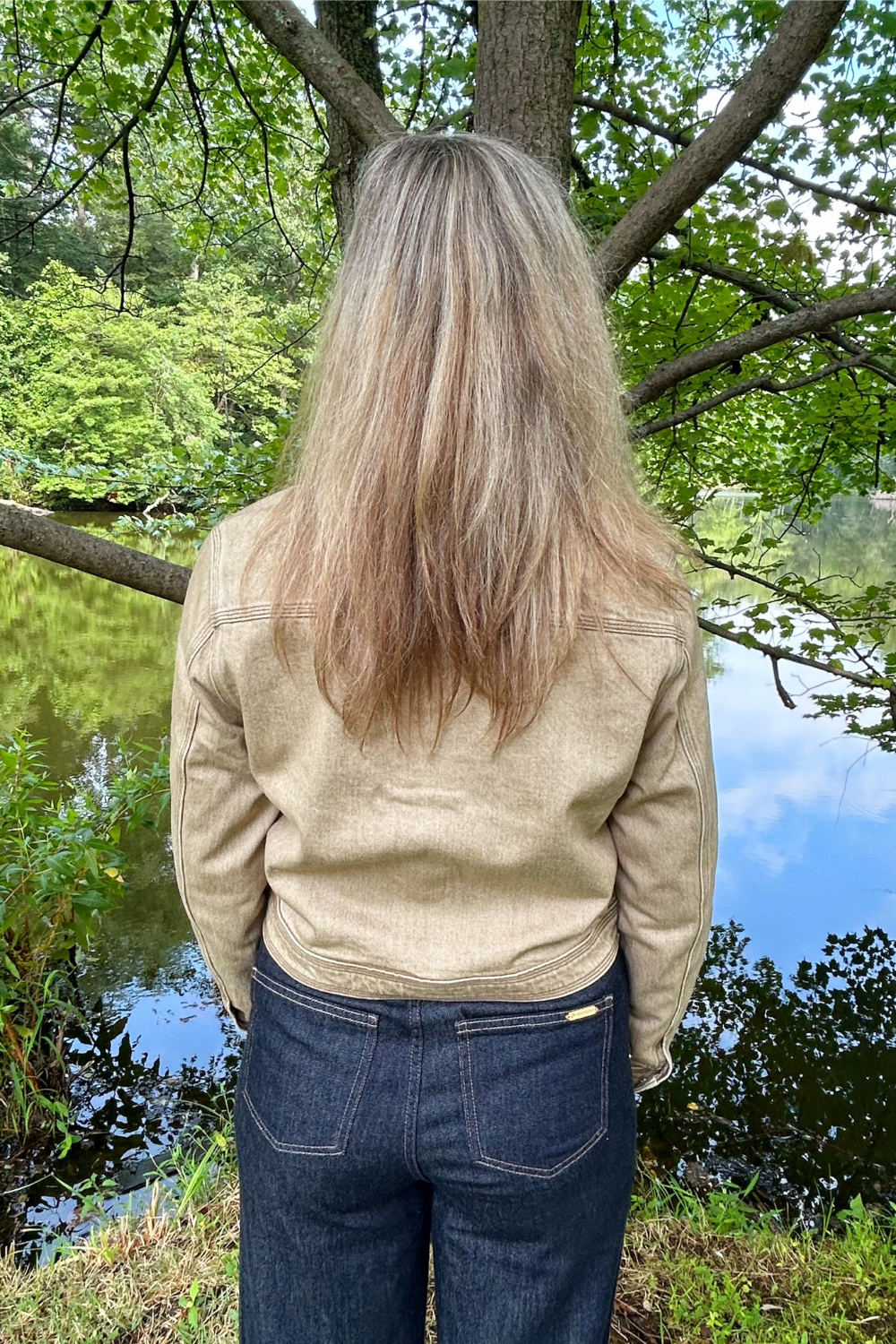 A person viewed from behind wearing a light beige canvas jacket and dark blue jeans, standing outdoors in front of a tree by a lake.