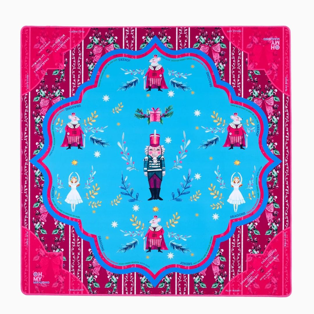 A square decorative textile or scarf laid flat on a white background, featuring a bright blue central field with nutcracker, ballerina, gift, and bird motifs, surrounded by a magenta border with floral and striped details.