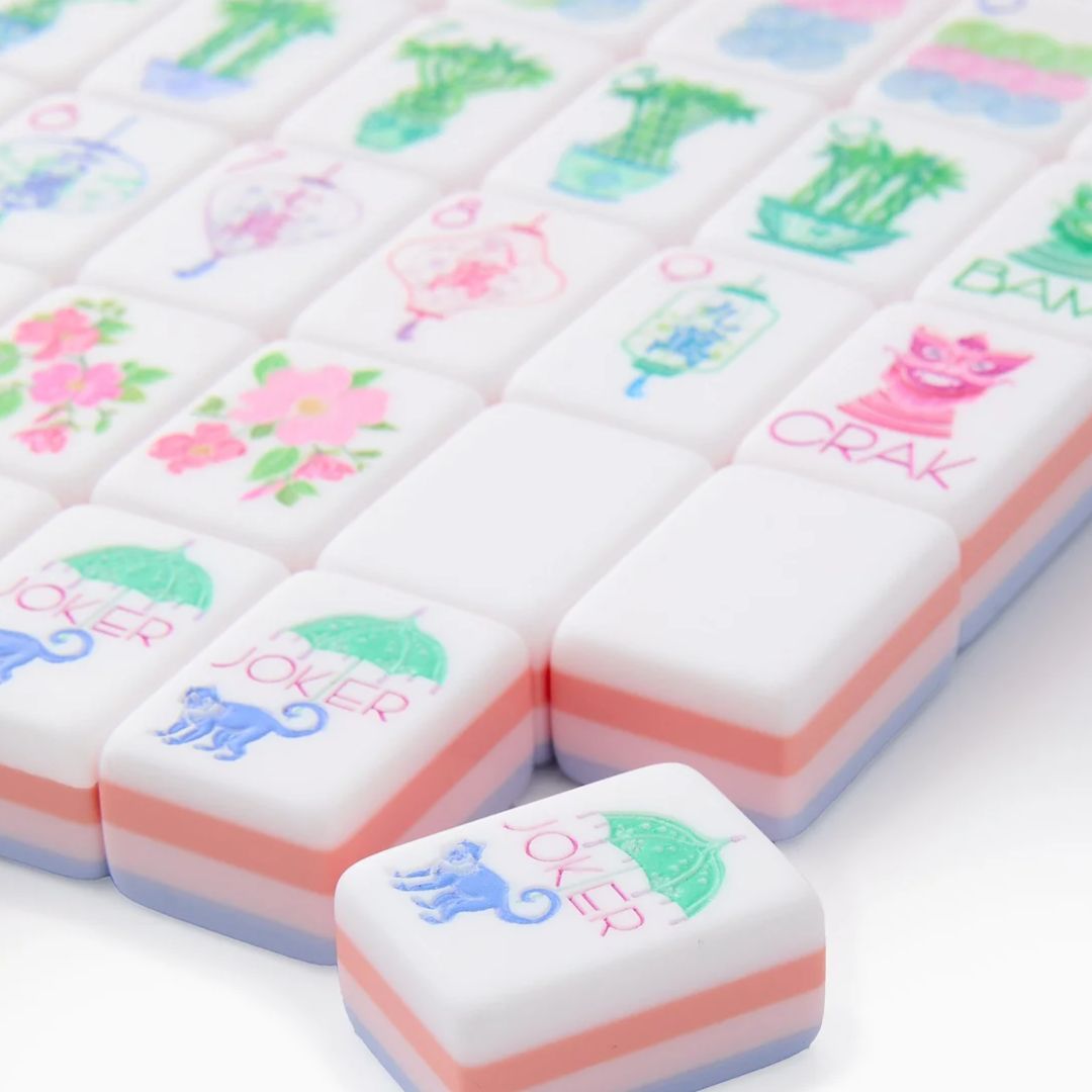 Close-up of white mahjong tiles with pastel pink and lavender sides, featuring colorful floral, bamboo, character, circle and joker designs arranged across a white surface.