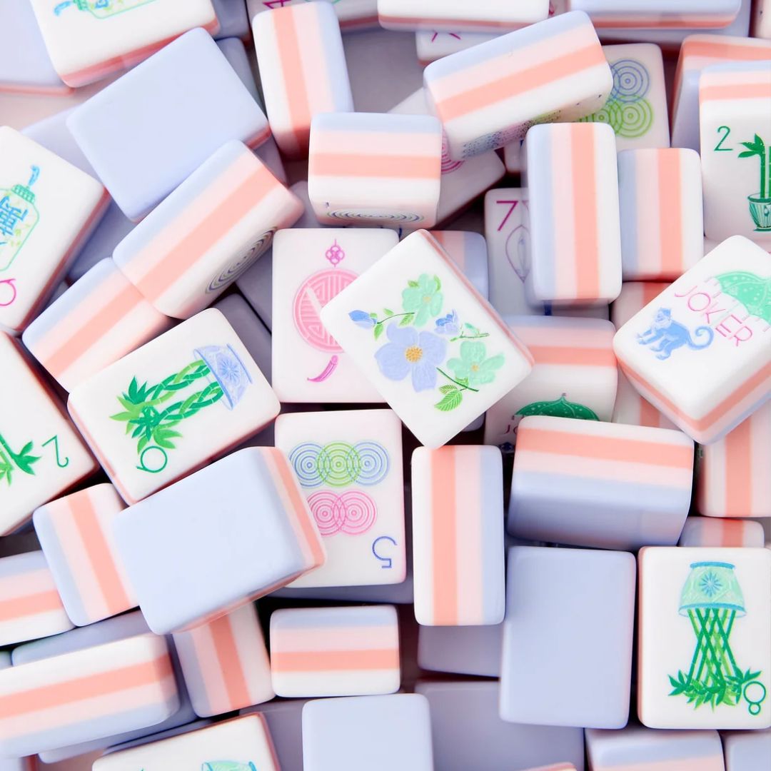 Close-up of white mahjong tiles with pastel pink and lavender sides, featuring colorful floral, bamboo, character, circle and joker designs arranged across a white surface.