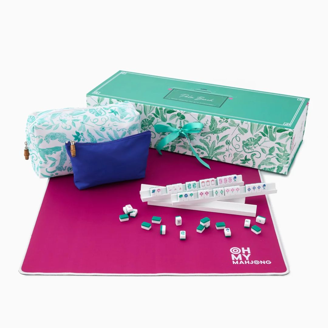 Mahjong set with a magenta game mat, white tile racks, scattered mahjong tiles, a blue pouch, and a patterned gift box on a white background.