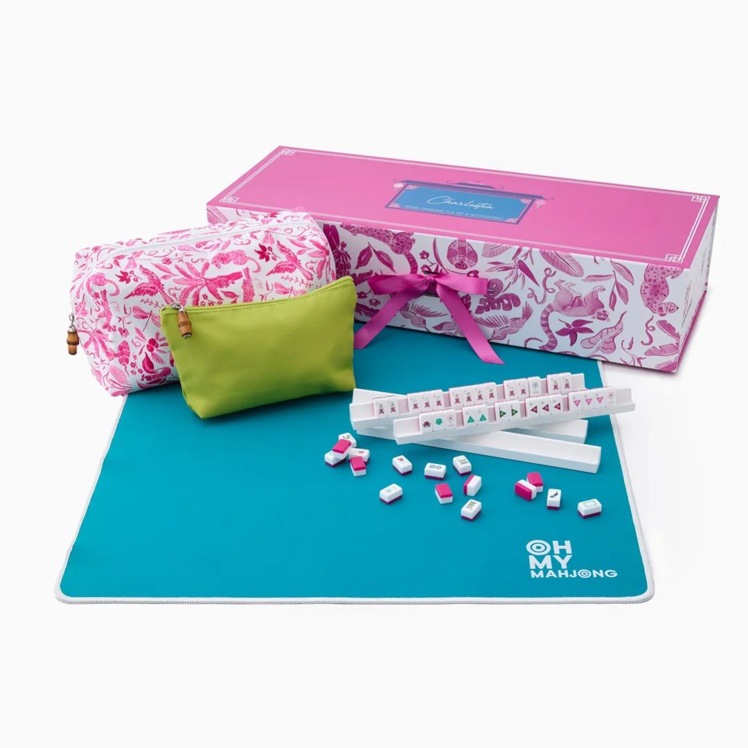 Oh My Mahjong set displayed on a teal mat with a pink patterned gift box, a pink-and-white patterned pouch, a small lime green pouch, and several white tiles with pink accents on a white background.