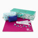 Mahjong set with a magenta game mat, white tile racks, scattered mahjong tiles, a blue pouch, and a patterned gift box on a white background.