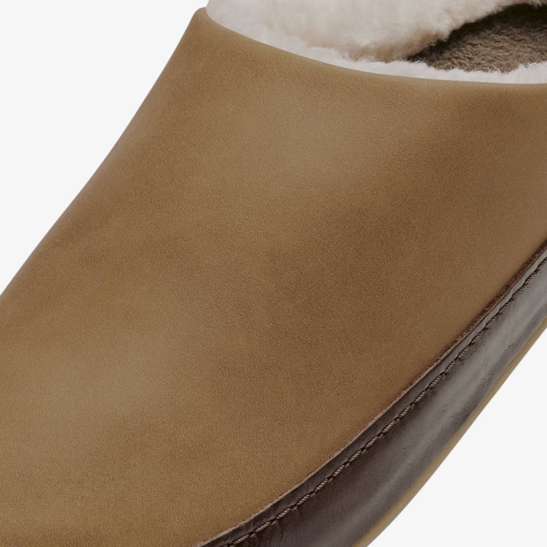 Close-up of a slip-on mule shoe in tan suede with a dark brown trim and white shearling lining.