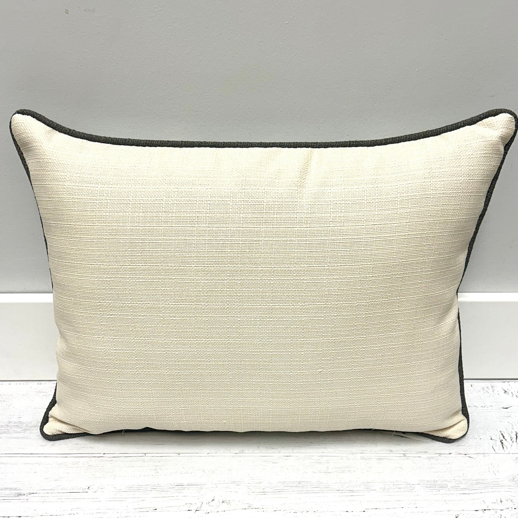 A rectangular lumbar throw pillow in textured cream fabric with dark charcoal gray piping, resting on a white wooden floor against a gray wall.