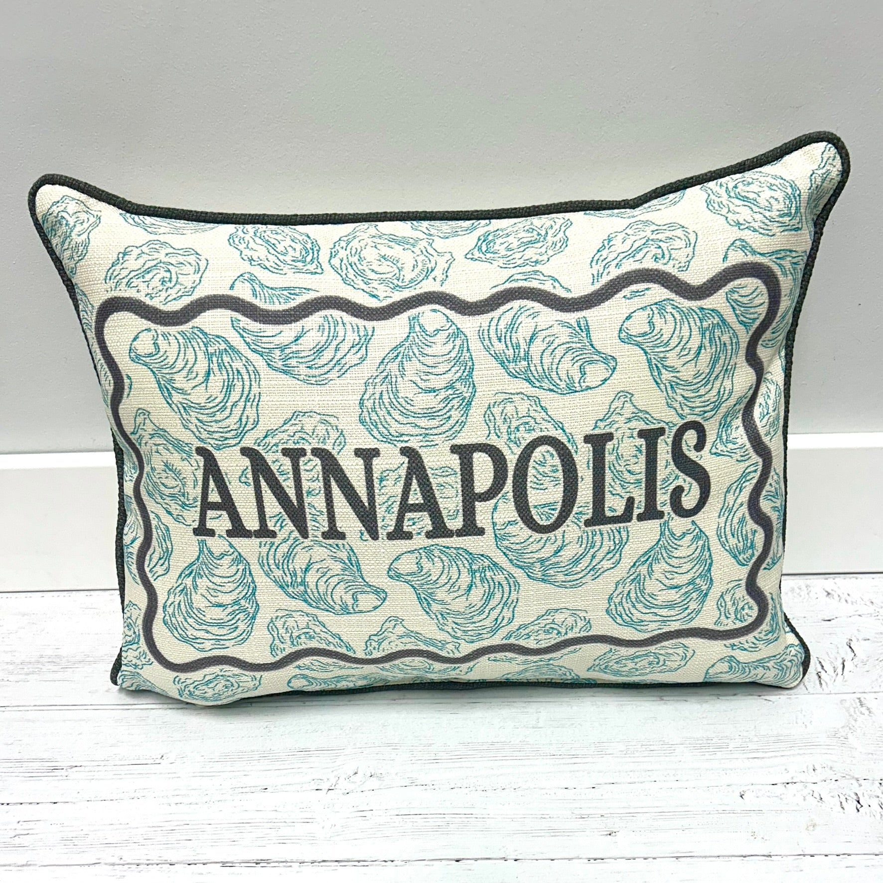 A rectangular decorative throw pillow with a cream background, teal oyster shell pattern, and black piping. The center features a black scalloped border with the word 'ANNAPOLIS' printed in black serif letters.