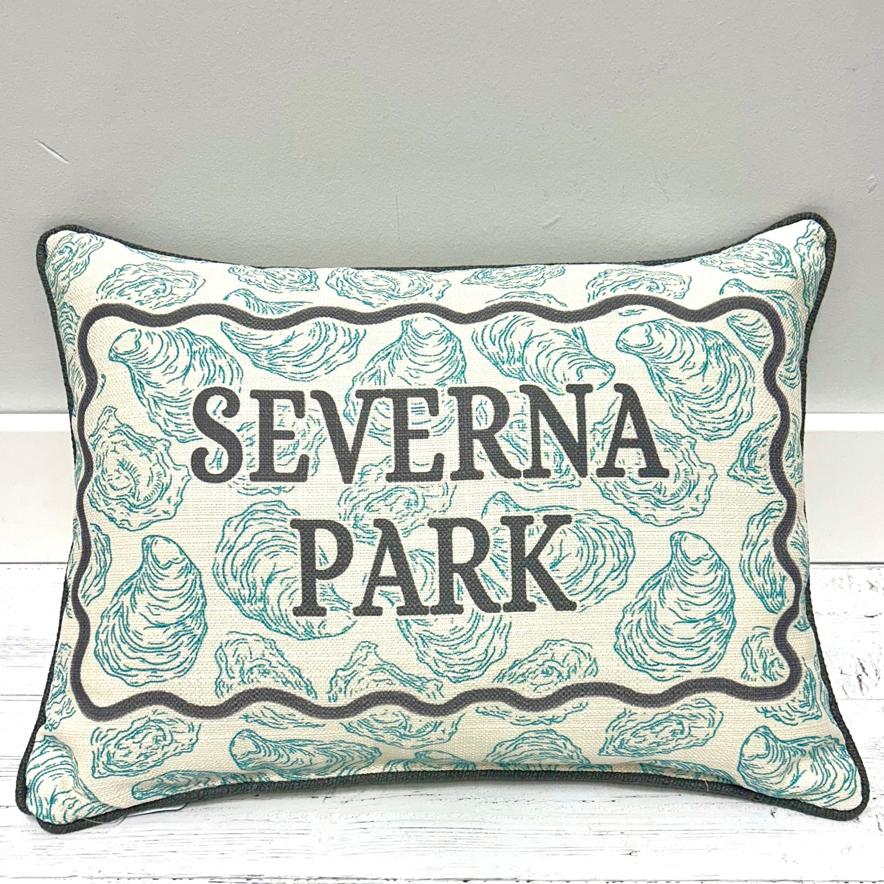 A square decorative throw pillow with a cream background, teal oyster shell pattern, and black text reading 'SEVERNA PARK' inside a wavy black border, shown leaning against a light gray wall on a white wood surface.