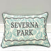 A square decorative throw pillow with a cream background, teal oyster shell pattern, and black text reading 'SEVERNA PARK' inside a wavy black border, shown leaning against a light gray wall on a white wood surface.