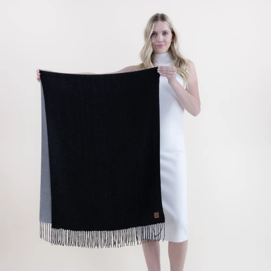 A woman in a white sleeveless dress holds up a large black knit scarf with fringe trim and a gray side panel against a plain light background.