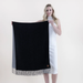 A woman in a white sleeveless dress holds up a large black knit scarf with fringe trim and a gray side panel against a plain light background.