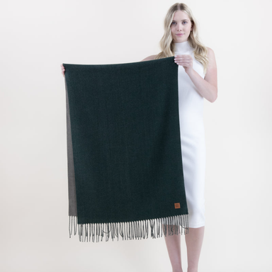 A woman in a white sleeveless dress holds up a large dark forest green woven scarf with fringe trim and a small brown leather logo patch, against a plain light beige studio background.