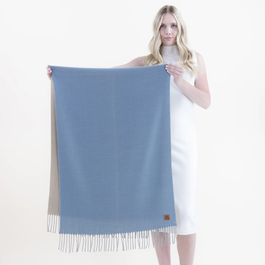 A woman in a white sleeveless dress holds up a large rectangular scarf or shawl with fringe. The front panel is dusty light blue, while the reverse side shown behind it is a warm beige.