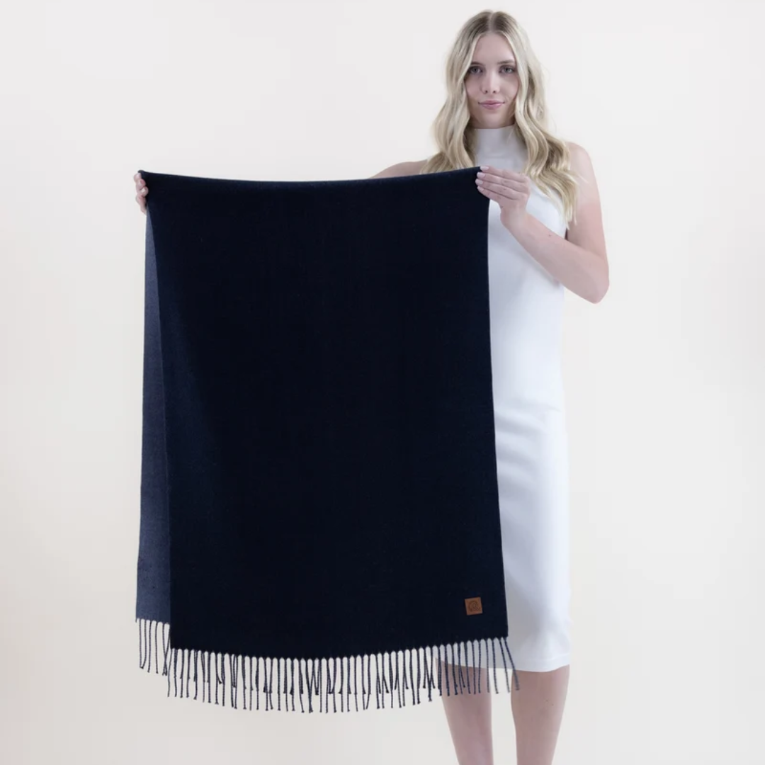 A woman in a white sleeveless dress holds up a large dark navy blue scarf with fringe trim against a plain light background.
