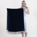A woman in a white sleeveless dress holds up a large dark navy blue scarf with fringe trim against a plain light background.