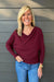 Woman wearing Six Fifty Anywhere Top In Burgundy with jeans on a dark wood background.