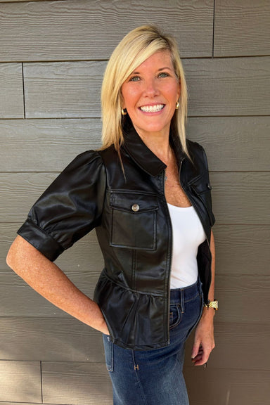 A woman wearing a color-blocked hoodie with a black hood and upper chest, dark gray middle chest, and heather gray sleeves and lower body, paired with blue jeans, standing against a plain light gray wall.