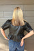 A woman viewed from behind wearing a black faux-leather peplum top with puff sleeves and blue jeans, standing against a gray wood-paneled wall.
