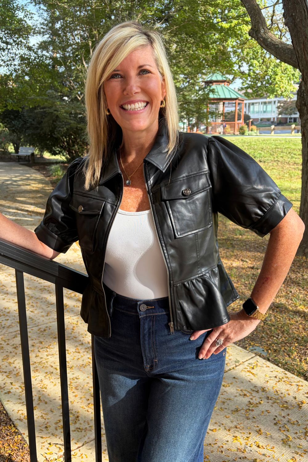 A smiling blonde woman wearing a black faux-leather moto jacket, white tank top, and blue jeans, standing outdoors on a paved path next to a black metal railing with trees in the background.