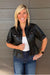 A smiling blonde woman wearing a black faux-leather cropped jacket with short puff sleeves, a white top, and blue jeans, standing against a gray wood-paneled wall.