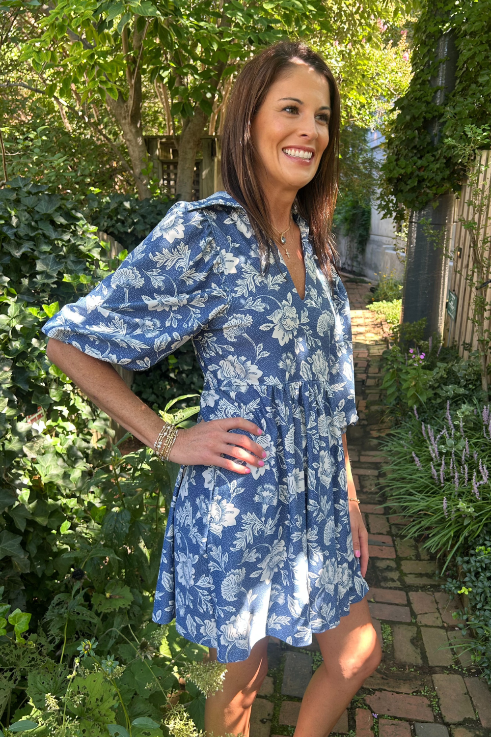 A woman wearing a blue and white floral print dress stands on a brick garden path surrounded by lush green foliage.