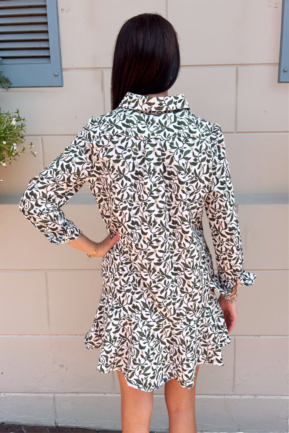 A woman viewed from behind wearing a long-sleeve ruffled shirt dress with an all-over abstract leaf-like print in green, white, and muted green, standing against a beige exterior wall.