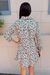 A woman viewed from behind wearing a long-sleeve ruffled shirt dress with an all-over abstract leaf-like print in green, white, and muted green, standing against a beige exterior wall.