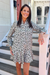 A woman wearing a long-sleeve mini dress with a green, white, and beige leafy pattern, standing on a  porch.