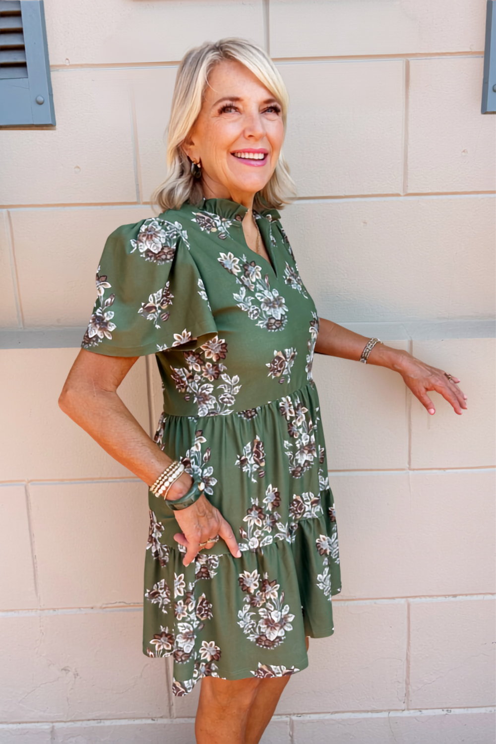 A woman with blonde hair wearing a green short-sleeve floral dress, posing against a beige brick wall.