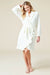 A woman modeling a white long-sleeve bathrobe with ruffled trim and a tie belt, standing against a plain beige studio background.