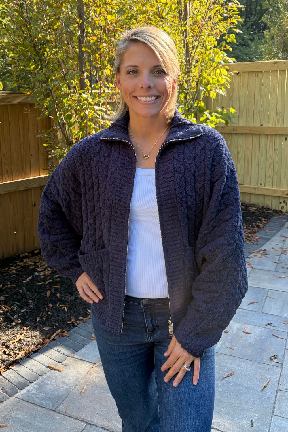 Woman wearing Softies Cable Knit Marshmallow Funnel Neck Double Zip Jacket Midnight with blue jeans on a patio that is fenced in.