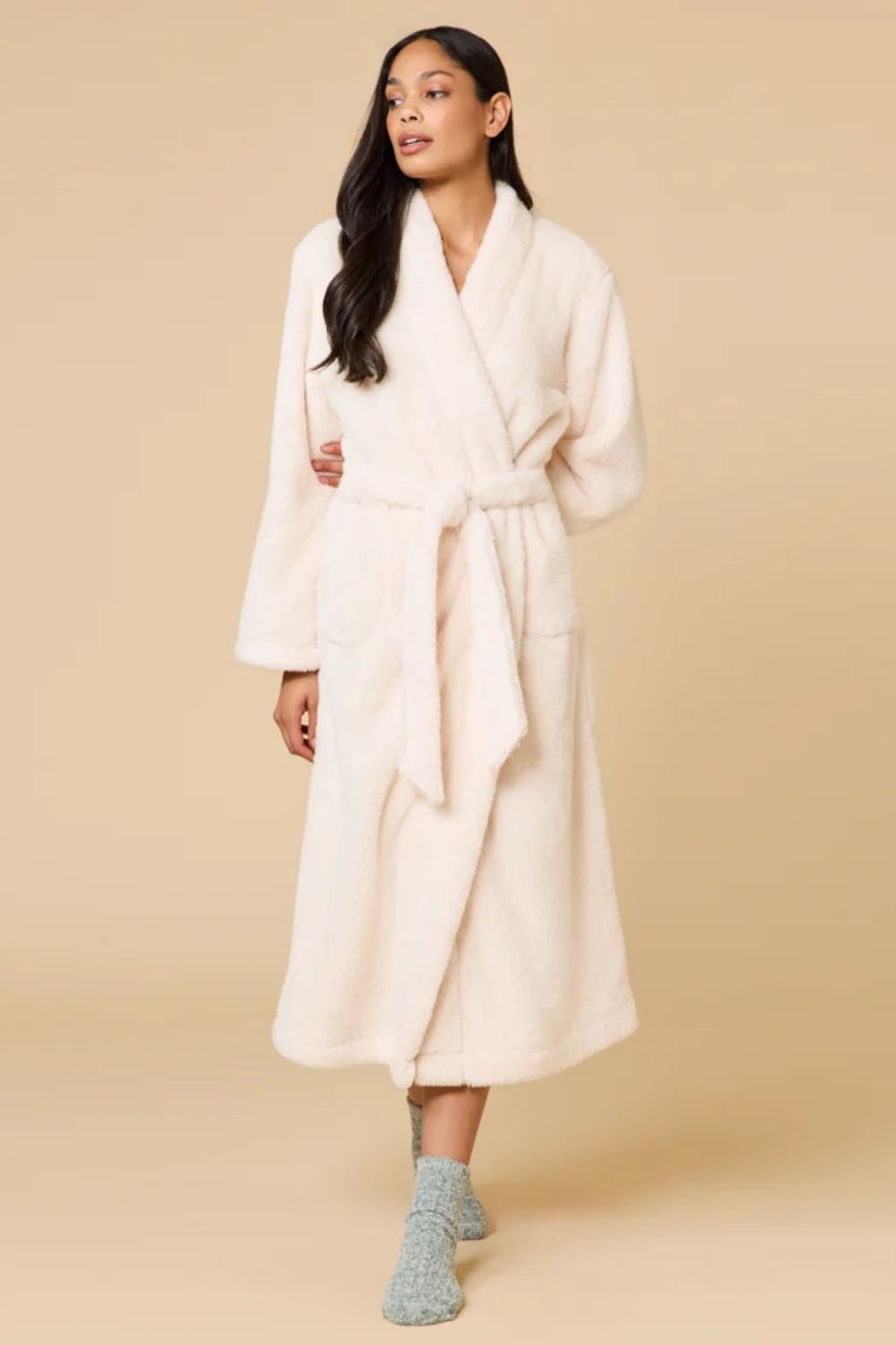 A woman modeling a plush, long cream-colored bathrobe with a tie belt, standing against a solid beige studio background.