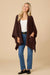 A woman modeling a dark brown open-front ribbed cardigan with pockets, worn over a white tank top and blue jeans, against a solid beige studio background.