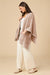 A woman modeling a light pink ribbed knit poncho cardigan over a white top and white wide-leg pants, standing against a solid beige studio background.