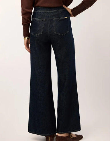 Close-up of a person wearing dark indigo denim jeans with a wide waistband  paired with a brown ribbed sweater against a light neutral background.