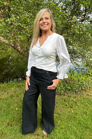 A woman standing outdoors on grass in front of trees, wearing a white long-sleeve blouse with ruffled cuffs and dark wide-leg pants.