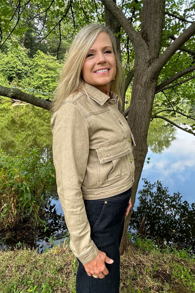 A smiling blonde woman wearing a light beige cropped denim jacket and dark jeans, standing outdoors next to a tree with green foliage and water in the background.
