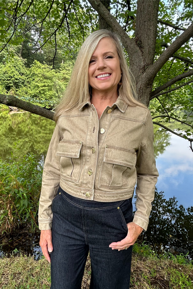 A smiling blonde woman wearing a light beige cropped utility jacket and dark jeans, standing outdoors in front of a large tree with green foliage and a blue sky visible.