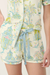A close-up of a person wearing a matching short-sleeve button-front top and shorts set with a tropical and architectural print in pastel blue, green, pink, and yellow on a cream background, tied with a light blue ribbon at the waist.