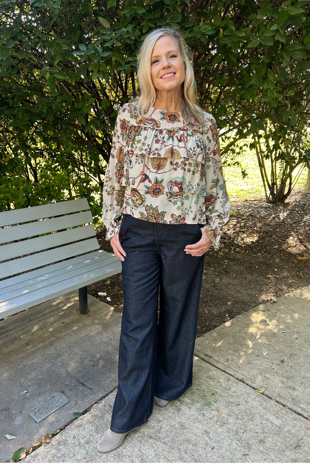 A woman stands outdoors on a concrete path in front of leafy green trees, wearing a cream blouse with a multicolor floral and paisley print, dark wide-leg jeans, and beige shoes.