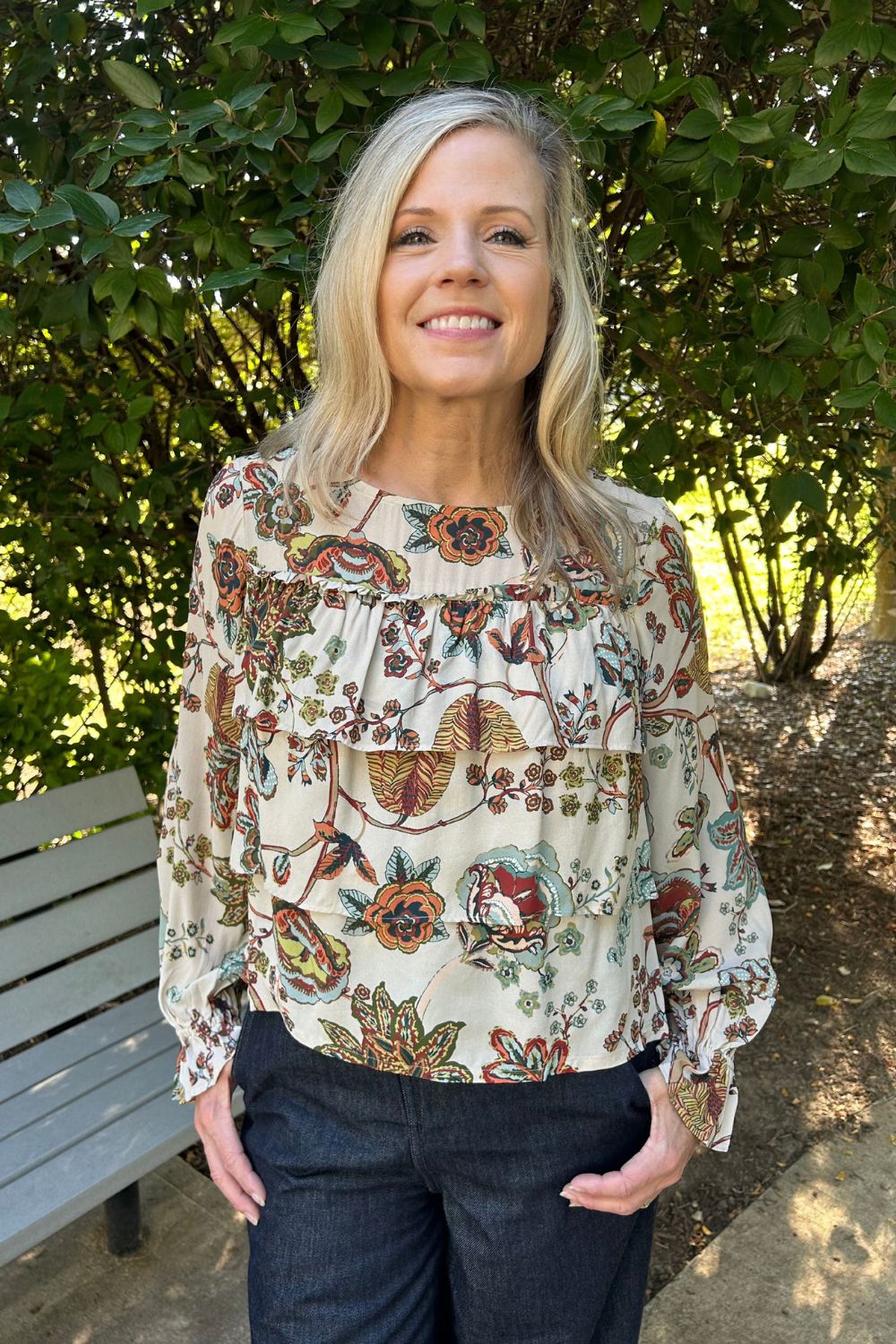 A smiling blonde woman wearing a cream-colored long-sleeve blouse with a detailed multicolor floral and botanical print, paired with dark blue jeans, standing outdoors in front of green bushes.