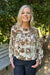 A smiling blonde woman wearing a cream-colored long-sleeve blouse with a detailed multicolor floral and botanical print, paired with dark blue jeans, standing outdoors in front of green bushes.