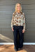 A woman stands against a gray wood-paneled wall wearing a long-sleeve cream blouse with a colorful floral and bird pattern, paired with dark wide-leg jeans.