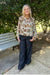 A woman stands outdoors on a concrete path in front of leafy green trees, wearing a cream blouse with a multicolor floral and paisley print, dark wide-leg jeans, and beige shoes.