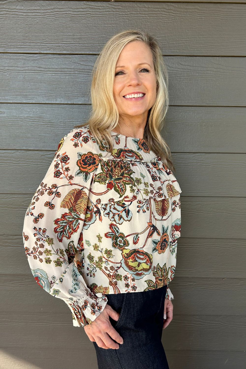 A smiling blonde woman wearing a cream-colored long-sleeve blouse with a detailed multicolor floral and botanical pattern, paired with dark jeans, standing against a gray wood-paneled wall.